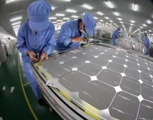 Workers in blue uniforms assemble a large solar cell module on a factory production line.