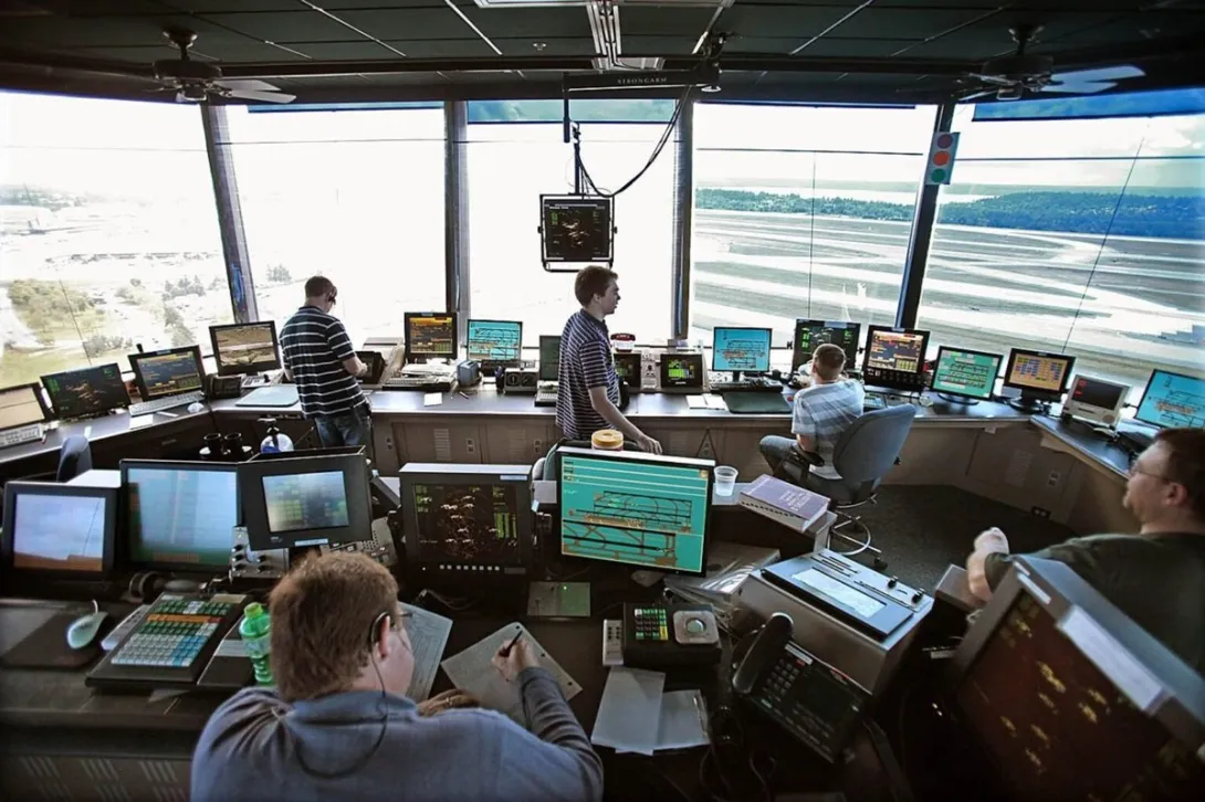 Air traffic controllers working at radar and flight displays inside an airport control tower overlooking the runways.