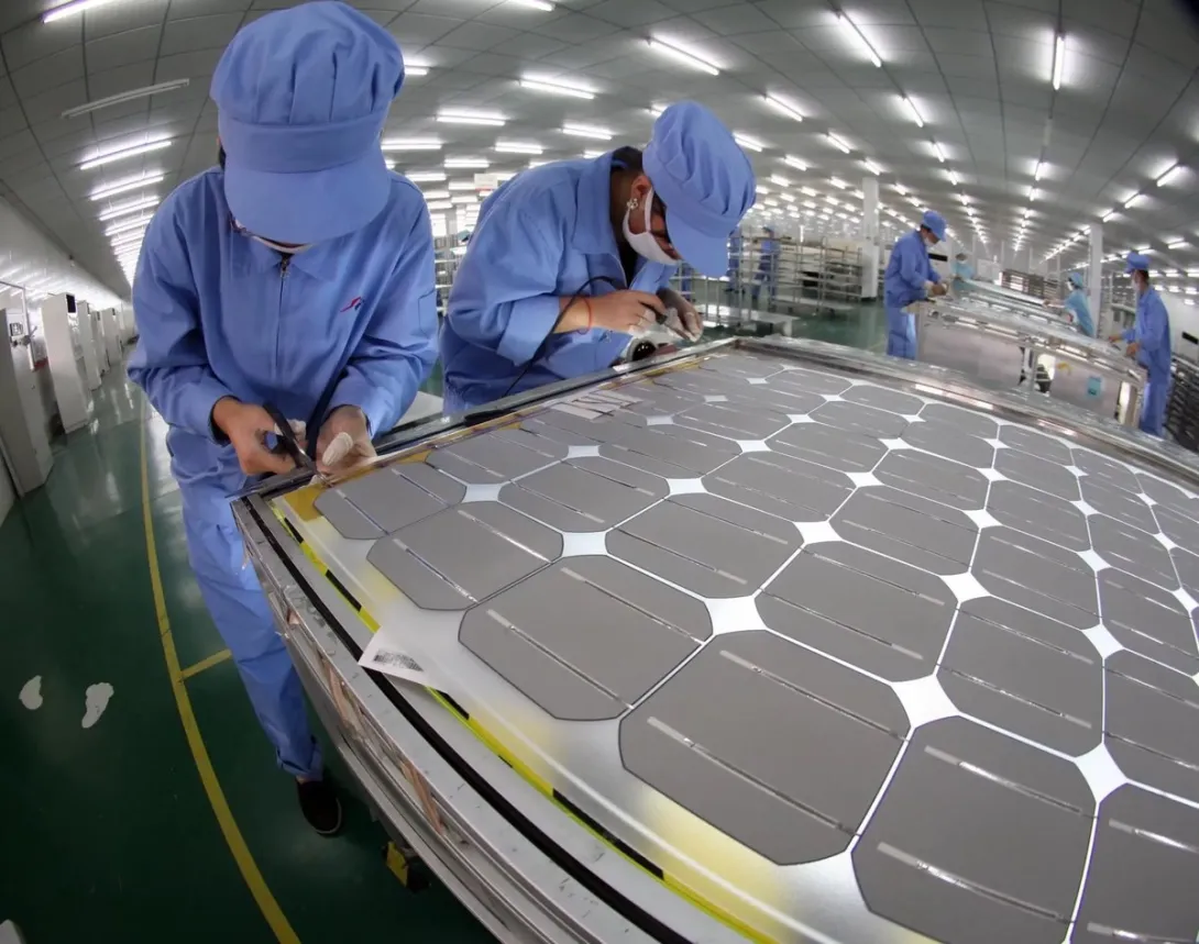 Workers in blue uniforms assemble a large solar cell module on a factory production line.